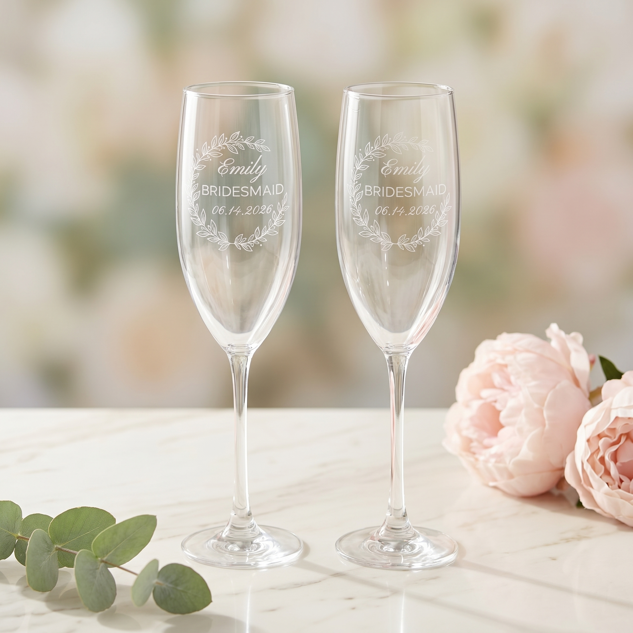 A matching pair of personalized champagne flutes engraved with the Eucalyptus Crescent Crest — one for the bride, one for her bridesmaid — styled together on white marble.