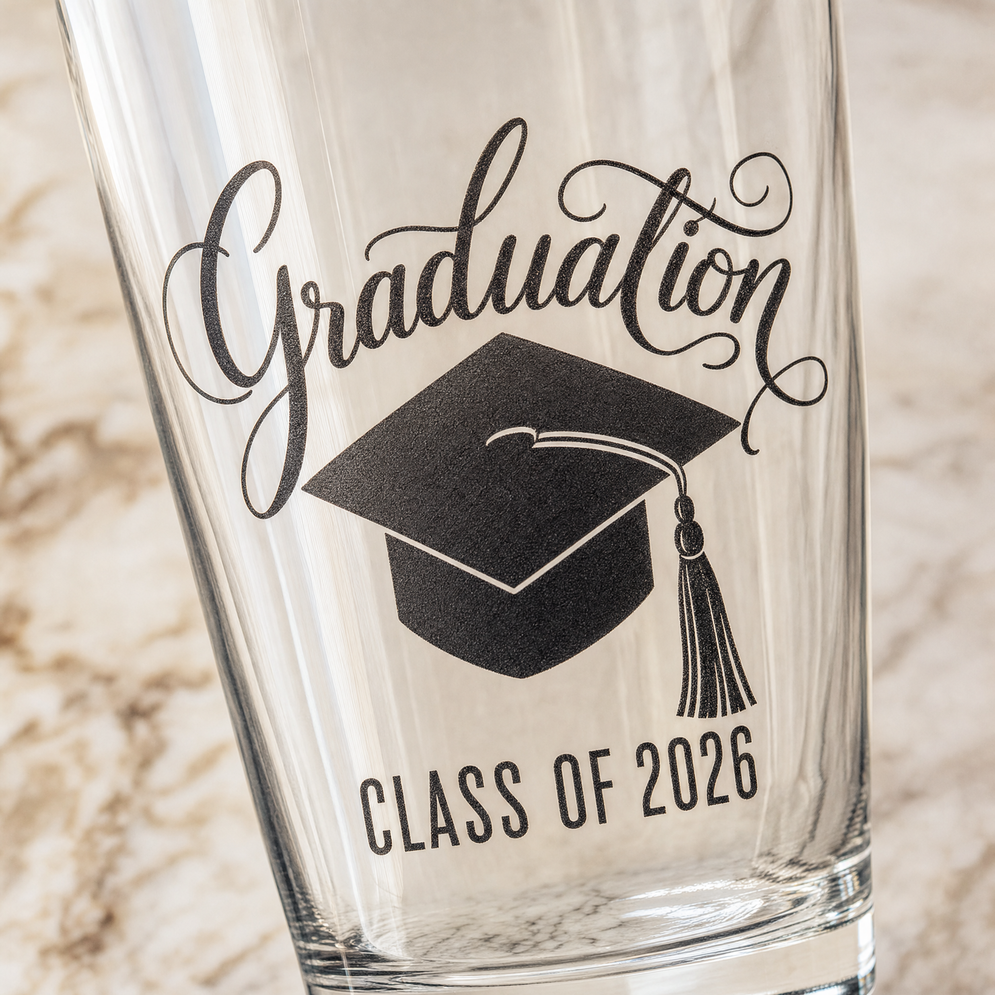 Macro closeup of the engraving — cursive 'Graduation' on top, black graduation cap silhouette in the middle, 'Class of 2026' in condensed sans-serif beneath.
