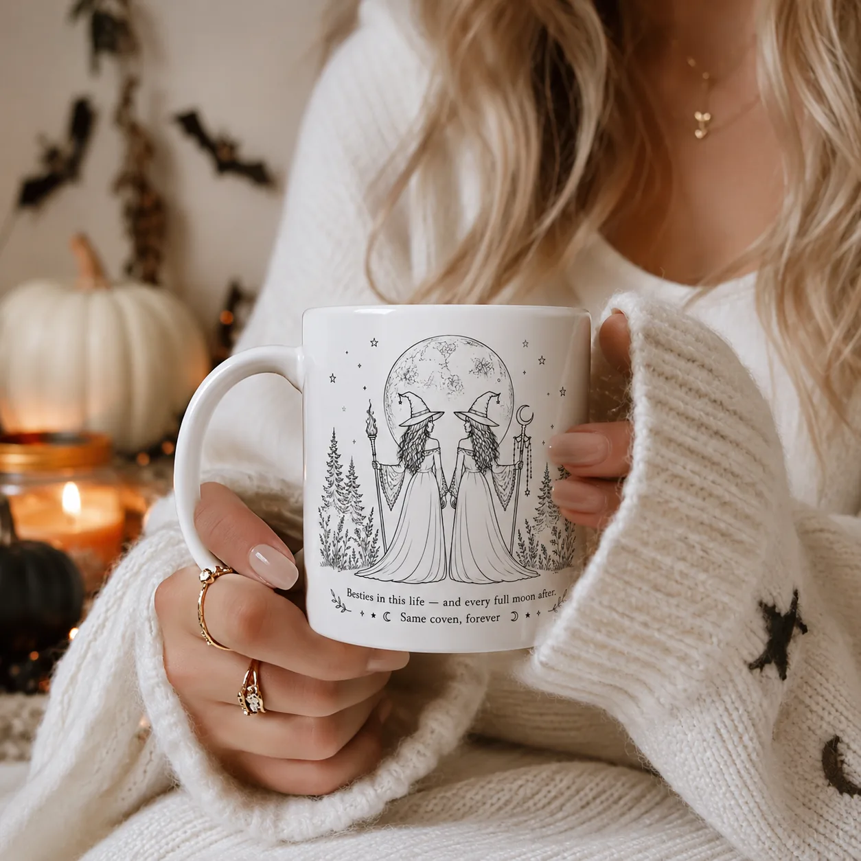 Halloween Bestie Gift: a personalized besties witch mug with two witches under a full moon, held by a woman in a cream sweater with candlelight and white pumpkins.